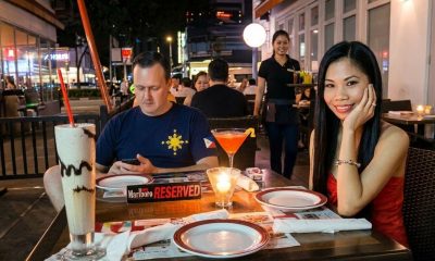 A man looking at his phone and a woman looking at the camera while seated have dinner outdoors in the Philippines.