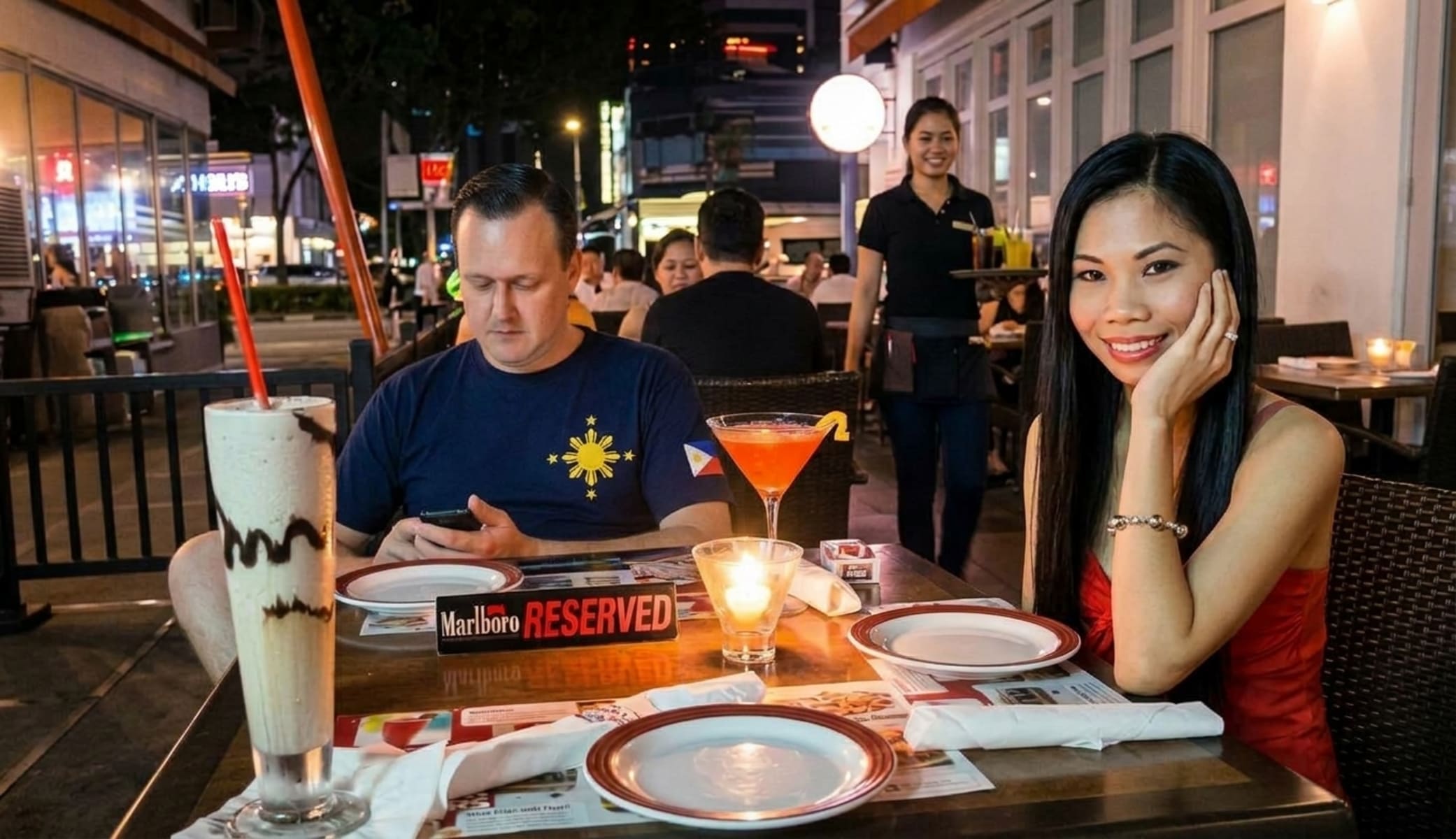 A man looking at his phone and a woman looking at the camera while seated have dinner outdoors in the Philippines.