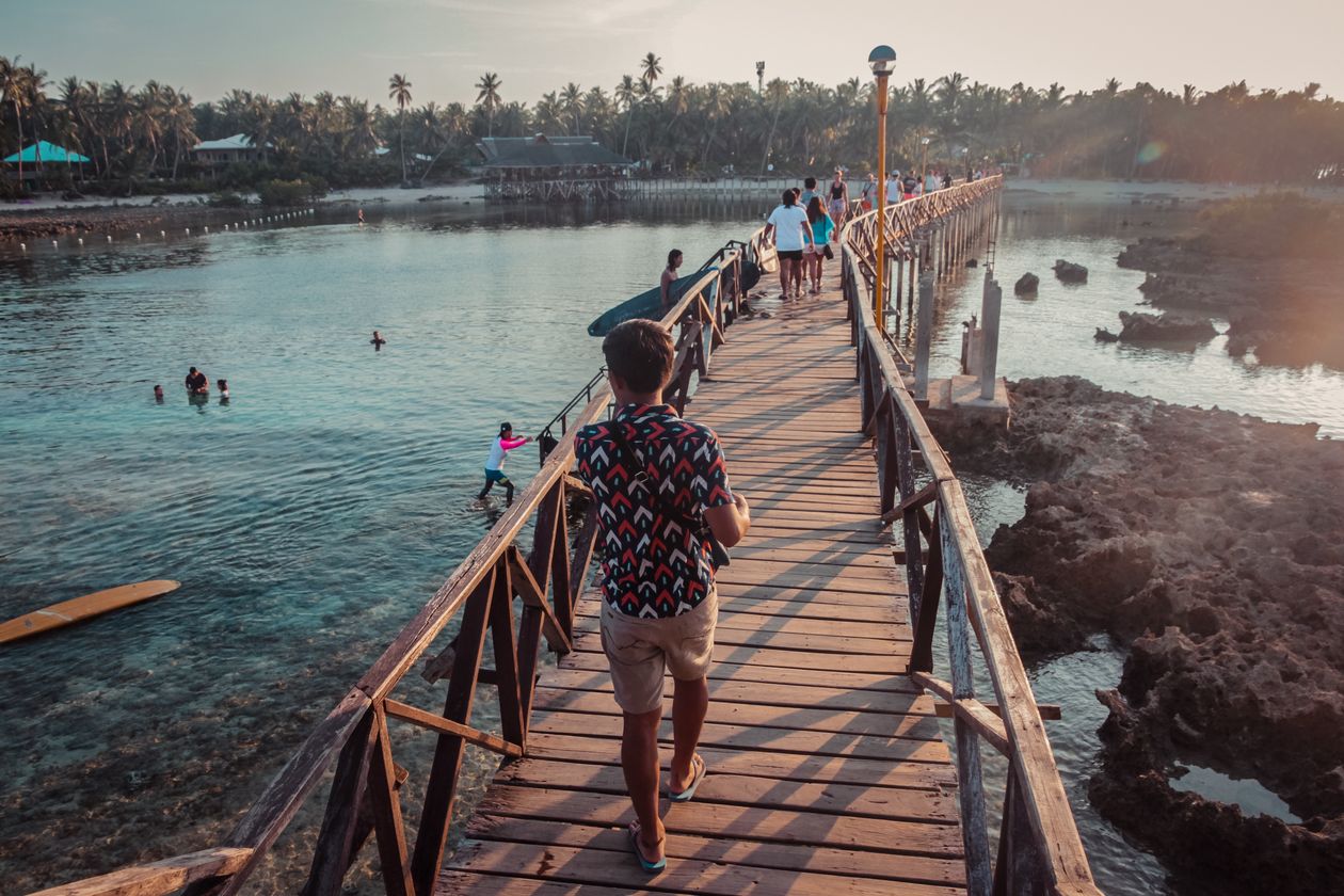 People walking along a wooden boardwalk over clear water at sunset in General Luna, Siargao