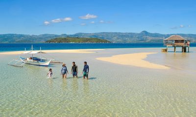 A small group of 4 people walking through shallow water on a sandbar with mountains in the background on a sunny day at the Manjuyod Sandbar, a day trip from Dumaguete, Philippines.
