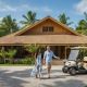 A man and woman walk along a gravel driveway in front of a tropical resort villa surrounded by palm trees, with a golf cart parked nearby.