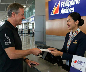 A man in a black SOLO Philippines shirt handing his ticket to a smiling airline agent at a Philippine Airlines check-in counter. Plan your solo trip with our 2-week itinerary.